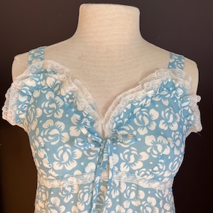 May include: A light blue floral print top with white flower designs. The top features a V-neckline with a delicate white lace trim and a matching bow. The straps are also adorned with lace.