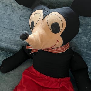 Puede incluir: Un mu&ntilde;eco de peluche vintage de Mickey Mouse. El mu&ntilde;eco presenta un cuerpo negro, grandes orejas negras, una cara beige con ojos negros y una bufanda a rayas rojas y blancas. Lleva pantalones cortos rojos.