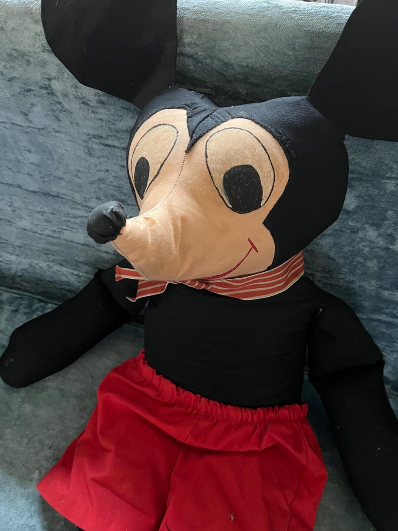Puede incluir: Un mu&ntilde;eco de peluche vintage de Mickey Mouse. El mu&ntilde;eco presenta un cuerpo negro, grandes orejas negras, una cara beige con ojos negros y una bufanda a rayas rojas y blancas. Lleva pantalones cortos rojos.