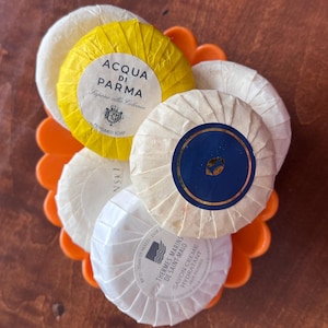 Op de afbeelding: Een verzameling ronde, afzonderlijk verpakte zepen in een feloranje, bloemvormige schaal. De zepen zijn wit en geel, met bedrukte etiketten. Eén label luidt "Acqua di Parma, Sapone alla Colonia, Perfumed Soap."