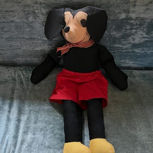 Puede incluir: Un mu&ntilde;eco de peluche grande de Mickey Mouse. El mu&ntilde;eco tiene un cuerpo negro, pantalones cortos rojos y pies amarillos. Tiene grandes orejas negras, una cara color canela y una bufanda a rayas rojas y blancas alrededor del cuello.