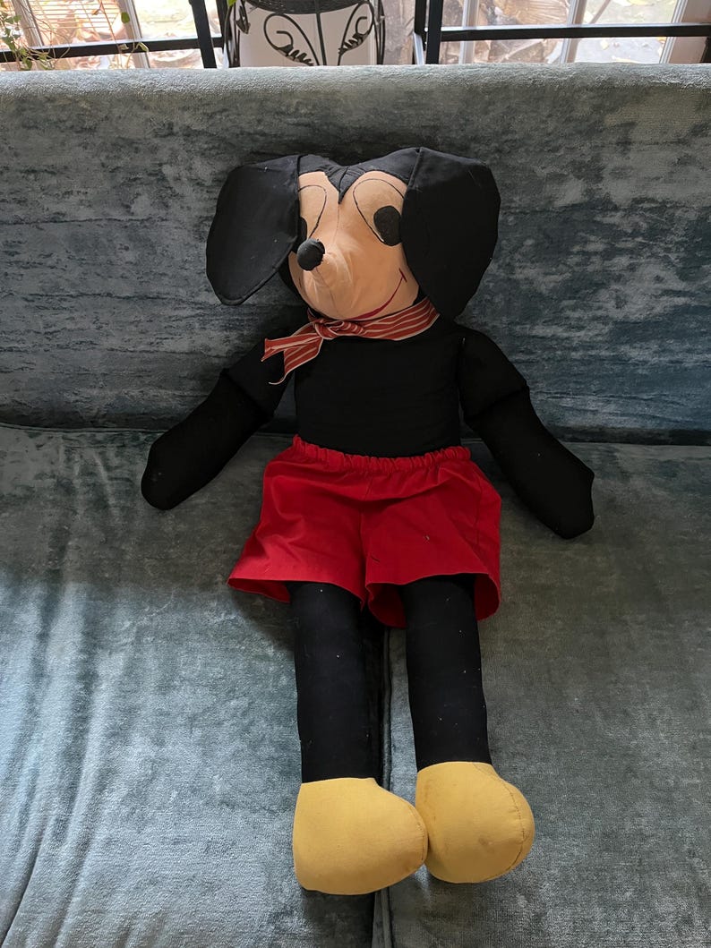 Puede incluir: Un mu&ntilde;eco de peluche grande de Mickey Mouse. El mu&ntilde;eco tiene un cuerpo negro, pantalones cortos rojos y pies amarillos. Tiene grandes orejas negras, una cara color canela y una bufanda a rayas rojas y blancas alrededor del cuello.