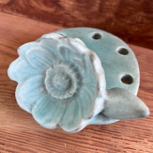 May include: A light blue ceramic flower bud vase with five holes for stems. The vase is shaped like a flower with a single large petal.
