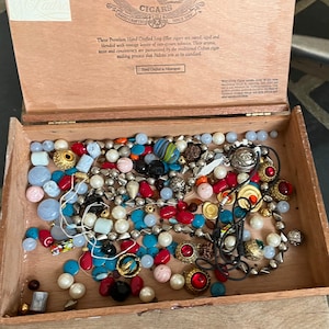 May include: A wooden box filled with a variety of colorful beads, including red, blue, white, gold, and black. The beads are arranged in a random pattern and some are strung together on a string. The box is made of wood and has a brown finish.