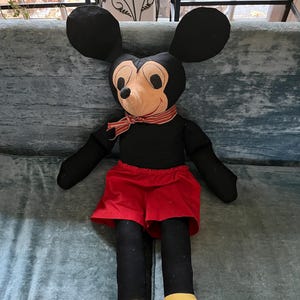 Puede incluir: Un mu&ntilde;eco de peluche grande de Mickey Mouse. El mu&ntilde;eco tiene un cuerpo negro, orejas grandes negras y una cara color canela con una bufanda a rayas rojas y blancas. Lleva pantalones cortos rojos y zapatos amarillos. El mu&ntilde;eco est&aacute; sentado sobre una superficie azul-verdosa.