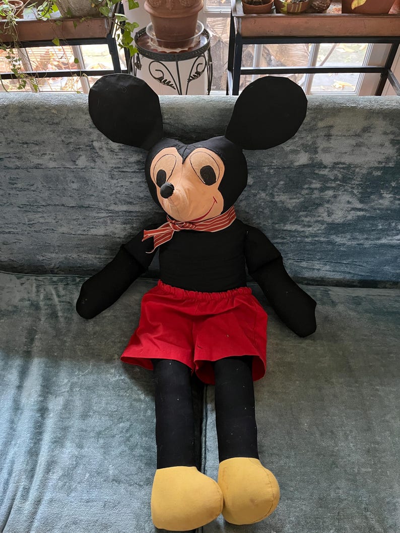 Puede incluir: Un mu&ntilde;eco de peluche grande de Mickey Mouse. El mu&ntilde;eco tiene un cuerpo negro, orejas grandes negras y una cara color canela con una bufanda a rayas rojas y blancas. Lleva pantalones cortos rojos y zapatos amarillos. El mu&ntilde;eco est&aacute; sentado sobre una superficie azul-verdosa.