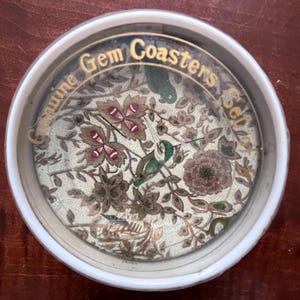 Op de afbeelding: Een set onderzetters met een bloemenontwerp en een kolibrie. De onderzetters zitten in een witte houder met gouden letters met de tekst "Genuine Gem Coasters Set". De onderzetters hebben een crèmekleurige achtergrond met bruine, groene en rode bloemen- en vogeldetails.