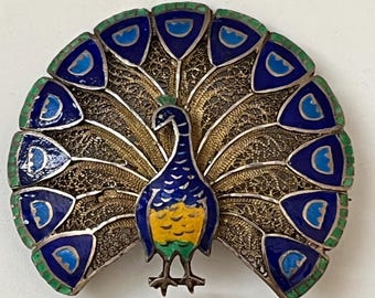 May include: A silver peacock brooch with blue, green, and yellow enamel. The peacock's tail feathers are made of filigree and are spread out in a fan shape.