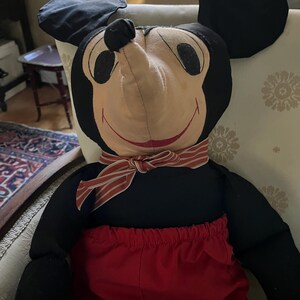 Puede incluir: Un mu&ntilde;eco de peluche vintage de Mickey Mouse. El mu&ntilde;eco presenta una cabeza y orejas negras, una cara bronceada, pantalones cortos rojos y una pajarita a rayas. El mu&ntilde;eco est&aacute; sentado en una silla estampada.