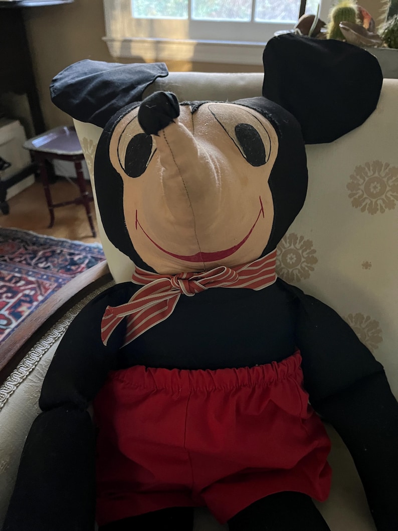 Puede incluir: Un mu&ntilde;eco de peluche vintage de Mickey Mouse. El mu&ntilde;eco presenta una cabeza y orejas negras, una cara bronceada, pantalones cortos rojos y una pajarita a rayas. El mu&ntilde;eco est&aacute; sentado en una silla estampada.