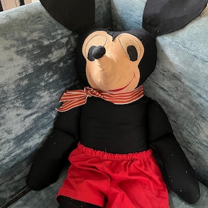 Puede incluir: Un mu&ntilde;eco de peluche vintage de Mickey Mouse con grandes orejas negras, una cara color canela y pantalones cortos rojos. El mu&ntilde;eco tiene una bufanda a rayas rojas y blancas alrededor del cuello y est&aacute; sentado sobre una superficie tapizada azul.