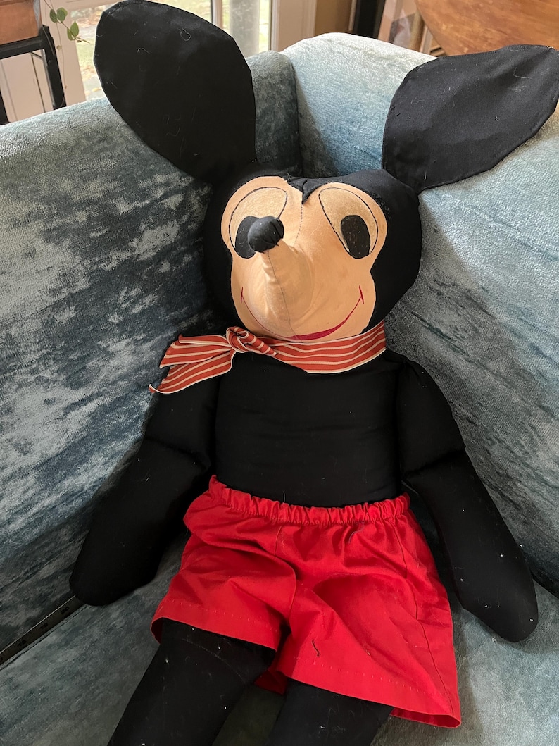 Puede incluir: Un mu&ntilde;eco de peluche vintage de Mickey Mouse con grandes orejas negras, una cara color canela y pantalones cortos rojos. El mu&ntilde;eco tiene una bufanda a rayas rojas y blancas alrededor del cuello y est&aacute; sentado sobre una superficie tapizada azul.