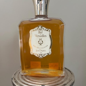 Könnte beinhalten: Eine durchsichtige Glasflasche mit Bal a Versailles Eau de Cologne von Jean Desprez Paris. Die Flasche hat ein goldenes Etikett mit einem dekorativen Rand und dem Text "Bal a Versailles Eau de Cologne Jean Desprez Paris". Die Flasche steht auf einem silbernen Ständer.