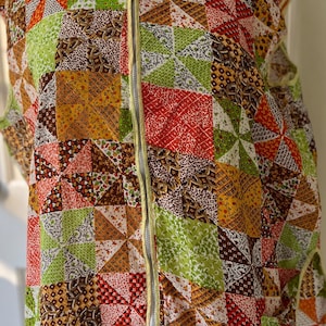 May include: A sleeveless, quilted vest featuring a patchwork design in various colors including green, orange, red, and brown. The vest has a full-length zipper and a yellow trim. The fabric is patterned with geometric shapes and floral motifs.