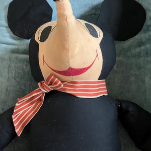 Puede incluir: Mu&ntilde;eco de peluche vintage de Mickey Mouse, con cuerpo negro, grandes orejas negras y cara beige. El mu&ntilde;eco tiene una bufanda a rayas rojas y blancas alrededor del cuello y una boca roja sonriente. Un personaje cl&aacute;sico de Disney.