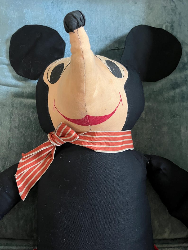 Puede incluir: Mu&ntilde;eco de peluche vintage de Mickey Mouse, con cuerpo negro, grandes orejas negras y cara beige. El mu&ntilde;eco tiene una bufanda a rayas rojas y blancas alrededor del cuello y una boca roja sonriente. Un personaje cl&aacute;sico de Disney.