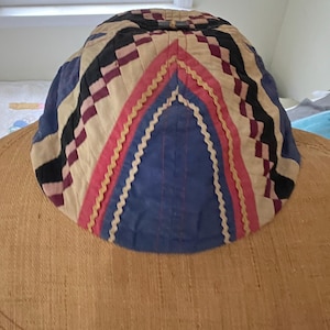 May include: A colorful, quilted cap with a geometric design. The cap features a combination of blue, black, cream, and pink fabrics. The central design is a blue triangle with pink and yellow trim. The cap is likely handmade.