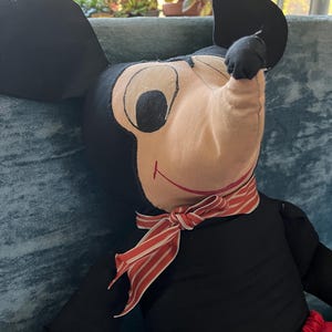 Puede incluir: Un mu&ntilde;eco de peluche vintage de Mickey Mouse. El mu&ntilde;eco tiene un cuerpo negro, orejas negras grandes y una cara beige con ojos negros. Lleva pantalones cortos rojos y una bufanda a rayas rojas y blancas. Un personaje cl&aacute;sico de Disney.
