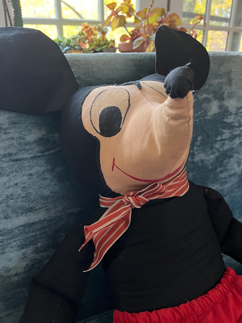 Puede incluir: Un mu&ntilde;eco de peluche vintage de Mickey Mouse. El mu&ntilde;eco tiene un cuerpo negro, orejas negras grandes y una cara beige con ojos negros. Lleva pantalones cortos rojos y una bufanda a rayas rojas y blancas. Un personaje cl&aacute;sico de Disney.