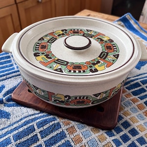 May include: A round, off-white ceramic casserole dish with a lid. The lid and the dish's rim feature a colorful, geometric pattern in orange, green, yellow, and brown. The dish has two side handles and sits on a wooden trivet.