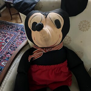 Puede incluir: Un mu&ntilde;eco de peluche grande de Mickey Mouse. El mu&ntilde;eco tiene orejas negras, un cuerpo negro y pantalones cortos rojos. Tiene una cara color crema con ojos negros y una bufanda a rayas rojas y blancas alrededor del cuello. El mu&ntilde;eco est&aacute; sentado en una silla.
