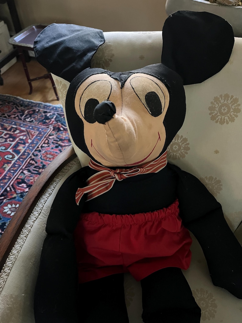 Puede incluir: Un mu&ntilde;eco de peluche grande de Mickey Mouse. El mu&ntilde;eco tiene orejas negras, un cuerpo negro y pantalones cortos rojos. Tiene una cara color crema con ojos negros y una bufanda a rayas rojas y blancas alrededor del cuello. El mu&ntilde;eco est&aacute; sentado en una silla.