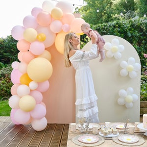 Pode incluir: Uma mulher com um vestido branco segura um bebê com um vestido rosa e branco no ar. Elas estão em pé na frente de um cenário com um arco de balões feito de balões rosa, amarelo e pêssego. O cenário é decorado com margaridas de papel branco.