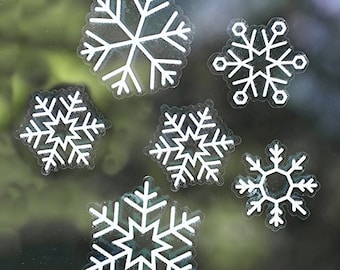 May include: A collection of six white snowflake stickers of varying sizes. Each snowflake features a detailed, symmetrical design with multiple points and intricate patterns. The stickers are displayed against a blurred background, suggesting they are adhered to a window.
