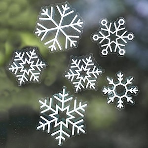Könnte beinhalten: Eine Sammlung von sechs weißen Schneeflocken-Aufklebern in verschiedenen Größen. Jede Schneeflocke hat ein detailliertes, symmetrisches Design mit mehreren Spitzen und komplizierten Mustern. Die Aufkleber werden vor einem verschwommenen Hintergrund gezeigt.