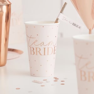 May include: Pink and gold paper cups with the text "Team Bride" printed on them. The cups are decorated with small gold dots.