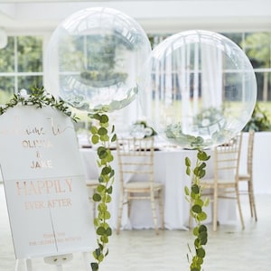 May include: Two clear balloons with greenery hanging from them. A white sign with the text "Welcome to Olivia & Jake Happily Ever After" is in the foreground.