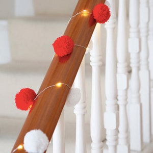 Puede incluir: Pompones blancos y rojos enhebrados en una cadena de luces blancas, decorando un pasamanos de madera en una escalera blanca.