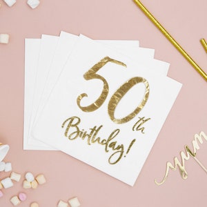 Può includere: Tovaglioli bianchi con scritta dorata in foglia che recita "50th Birthday!" Una cannuccia dorata glitterata, un topper per torta dorato glitterato che dice "yum" e marshmallow rosa e bianchi sono sparsi intorno ai tovaglioli.