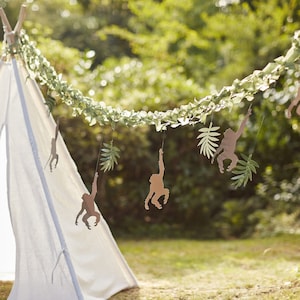 May include: A green garland with paper cutouts of monkeys hanging from it. The garland is strung between a white teepee tent and a green bush.