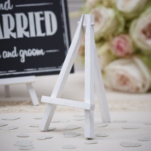 May include: White wooden easel stand for displaying small signs or photos. The easel is on a white surface with white paper hearts scattered around it.