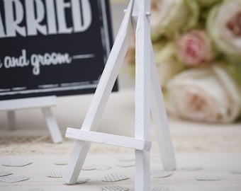 May include: White wooden easel stand for displaying small signs or photos. The easel is on a white surface with white paper hearts scattered around it.