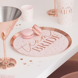 May include: Pink and rose gold paper plate with the text "Team Bride" in a script font. The plate has a white polka dot pattern and is decorated with two pink macarons.