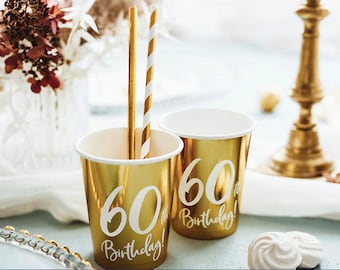 May include: Two gold paper cups with white lettering that says "60th Birthday!"  The cups are sitting on a white surface with a gold candlestick in the background.