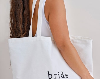 May include: A white tote bag with the word "bride" printed in black. The bag has two long straps and is made of a sturdy fabric. The bag is being held by a person.