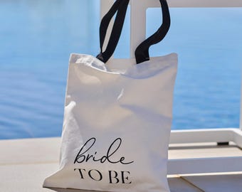 May include: A white canvas tote bag with black handles. The bag features the words "bride TO BE" in a stylish, handwritten font. The background shows a blue body of water and a white table.