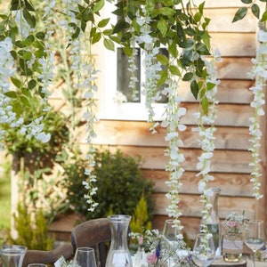 May include: White artificial wisteria flower garland hanging from a ceiling, with green leaves and white flowers. The garland is hanging over a table with glasses and a pitcher.