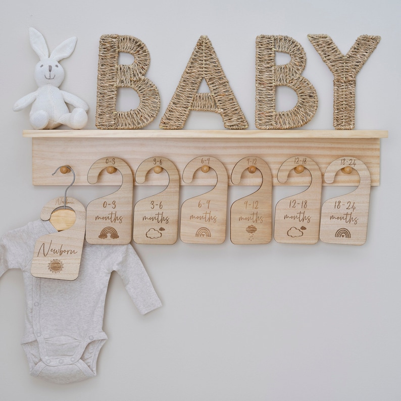 May include: Wooden hangers with engraved text indicating age ranges for a baby's clothing, such as "Newborn", "0-3 months", "3-6 months", and so on. The hangers are displayed on a wooden shelf with the word "BABY" spelled out in woven rope letters.