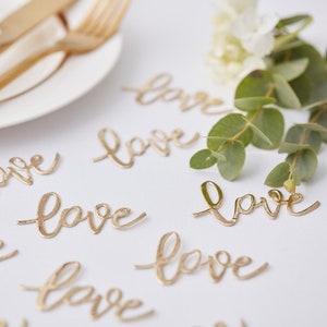 Peut inclure: Confettis dorés avec le mot "love" écrit sur chaque pièce. Les confettis sont éparpillés sur une surface blanche.