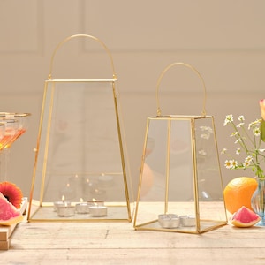 Op de afbeelding: Twee goudomrande glazen lantaarns met gebogen handvatten. De lantaarns van verschillende afmetingen bevatten brandende waxinelichtjes. De achtergrond omvat een tafel met fruit en bloemen.