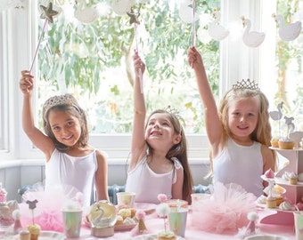 May include: Three girls wearing white tank tops and tulle skirts are sitting at a table decorated with pink and white swan-themed party supplies. They are holding up silver wands with star toppers. The table is set with pink plates, pink napkins, and pink cups. There are cupcakes and a tiered cake stand with cupcakes on it.