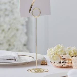 May include: Gold metal table number holder with a circular top and a spiral base. The holder is standing on a white tablecloth.