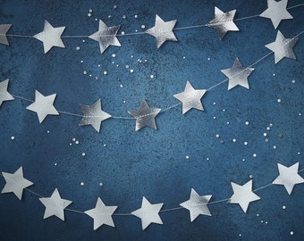 May include: A silver star garland hangs against a dark blue background. The garland is made of paper stars and is strung together with white thread. The background is speckled with silver glitter.