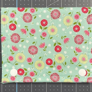 May include: A rectangular fabric item with a floral pattern in red, pink, and yellow on a light green background. The design features stylized flowers and small white blossoms. Two white snap closures are visible.
