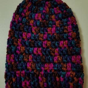 May include: A colourful, hand-crocheted beanie hat. The hat is made with a textured stitch and features a mix of purple, pink, teal, and orange yarns. This warm hat is ideal for colder weather and adds a bright touch to any outfit.