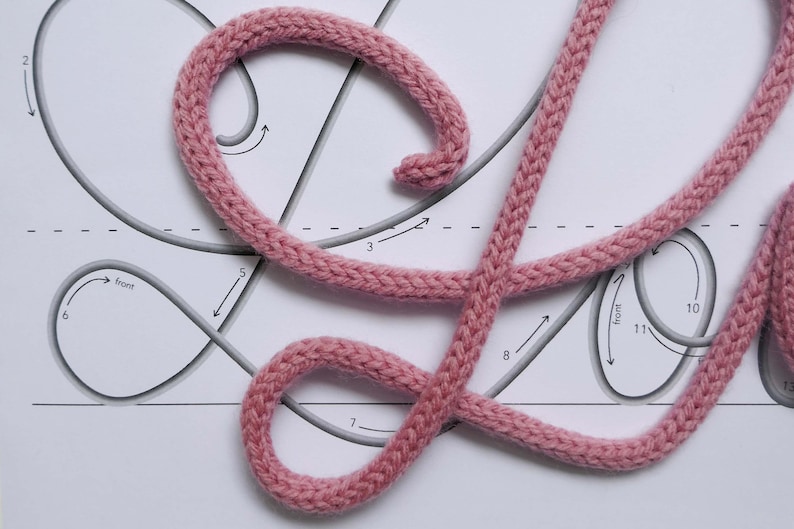 May include: A pink cord is laid out on a white surface, following a numbered pattern of lines and arrows. The cord is woven in a braided pattern.